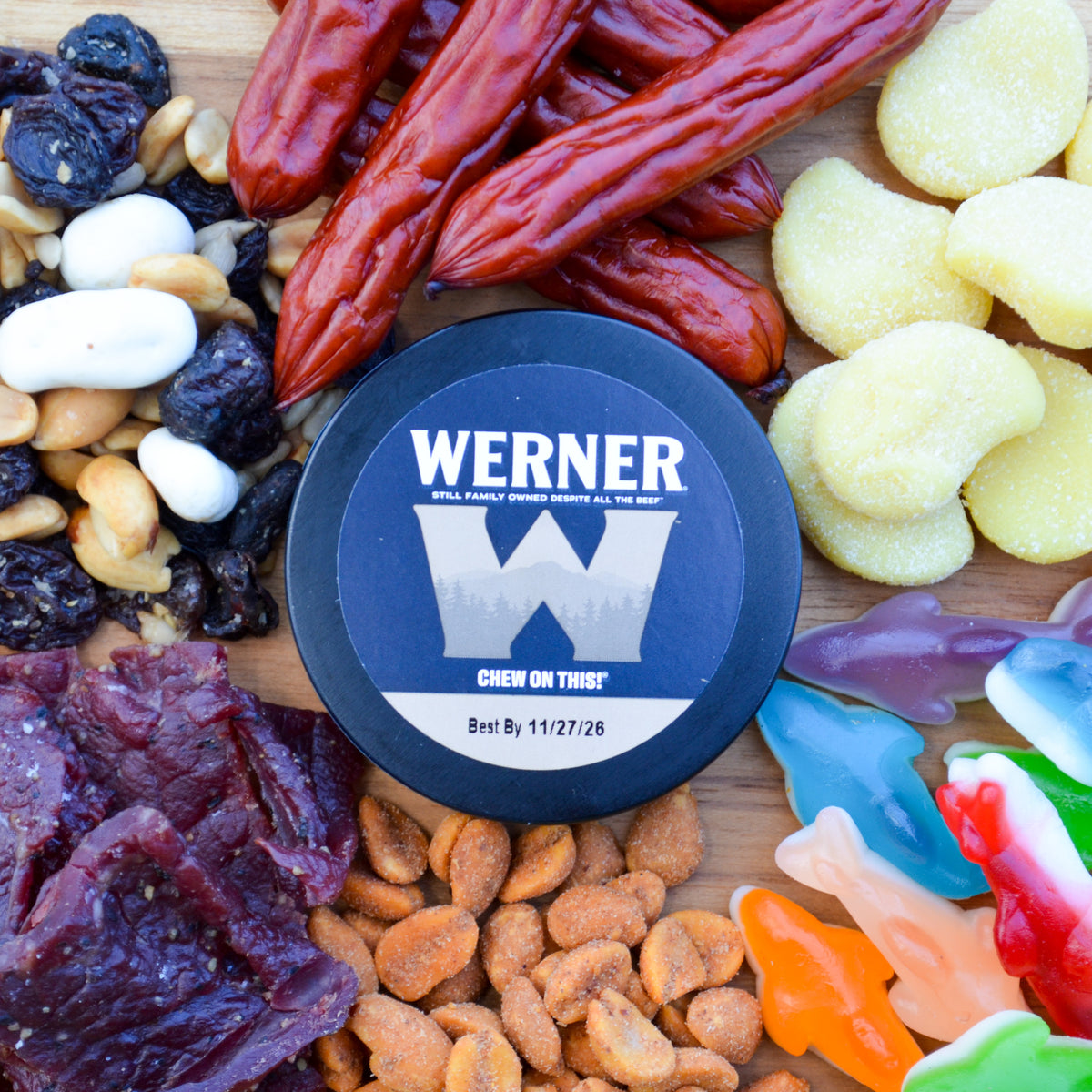 A round black container of Werner Chew on This! dog treats is centered on a wooden board surrounded by assorted snacks, including dried red chilies, yellow gummy candies, mixed nuts, dried fruit, and colorful gummy candies.