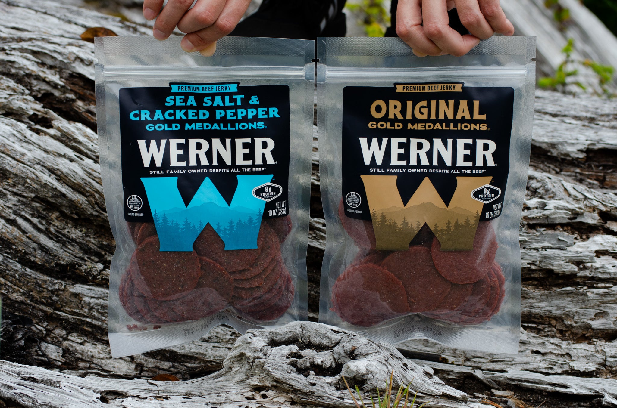 Two clear stand-up pouches of Werner meat snacks are held in front of a weathered log outdoors. The left pouch is labeled Sea Salt & Cracked Pepper Gold Medallions with a blue W logo, and the right pouch is labeled Original Gold Medallions with a gold W logo.
