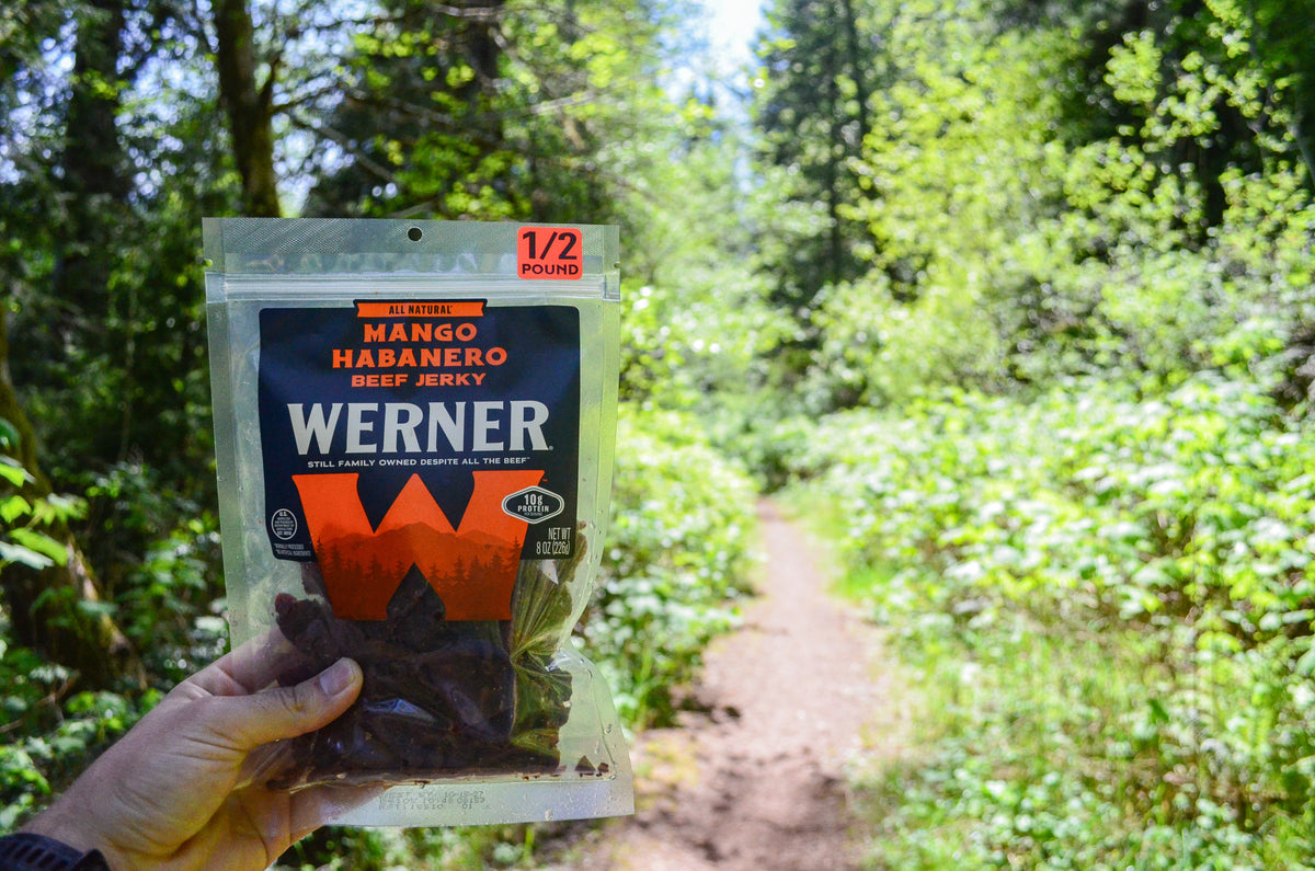 A hand holds a package of Werner Mango Habanero Beef Jerky in front of a blurred forest trail background.