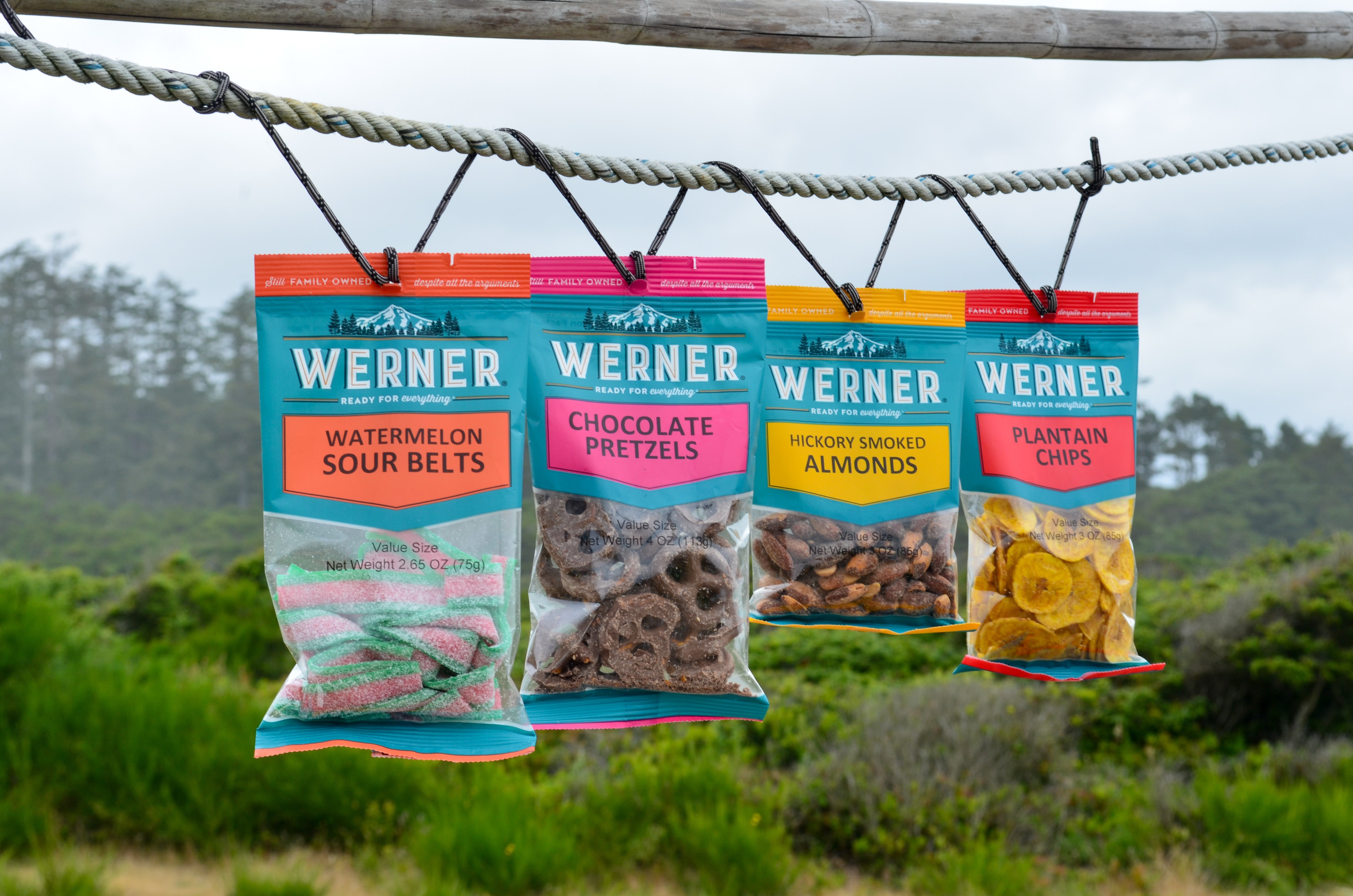 Four bags of Werner outdoor snacks hanging from a rope outdoors. The bags are for Watermelon Sour Belts, Chocolate Pretzels, Hickory Smoked Almonds, and Plantain Chips, with a blurred green forest background.